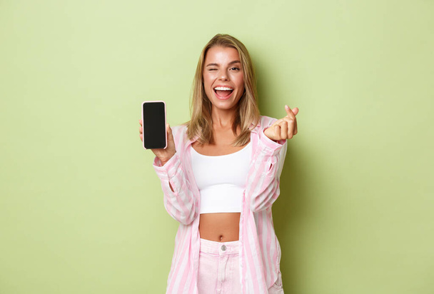 Image of stylish blond girl in pink shirt, showing heart sign and mobile phone screen, like something online, standing over green background - Fotó, kép