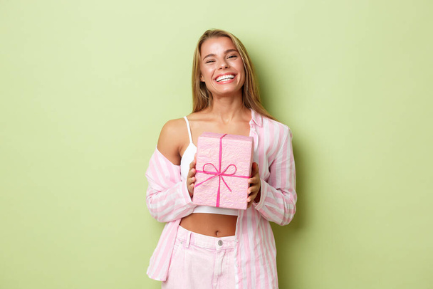 Imagen de la atractiva chica rubia celebrando las vacaciones, sonriendo feliz como de pie con caja de regalo envuelta en rosa sobre fondo verde - Foto, imagen