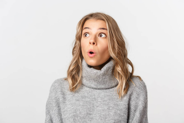 Close-up of pretty blond girl in grey sweater, looking at upper left corner with surprised face, gasping fascinated, standing over white background - Fotó, kép