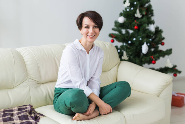 Счастливая молодая женщина на диване в гостиной с елкой. Концепция праздников. - Фото, изображение