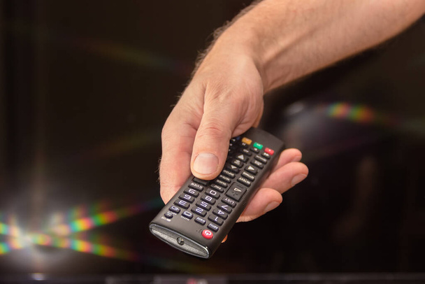 Телевизионный пульт дистанционного управления в руке человека на размытом темном фоне с лучами и вспышками света, крупным планом в избирательном фокусе - Фото, изображение