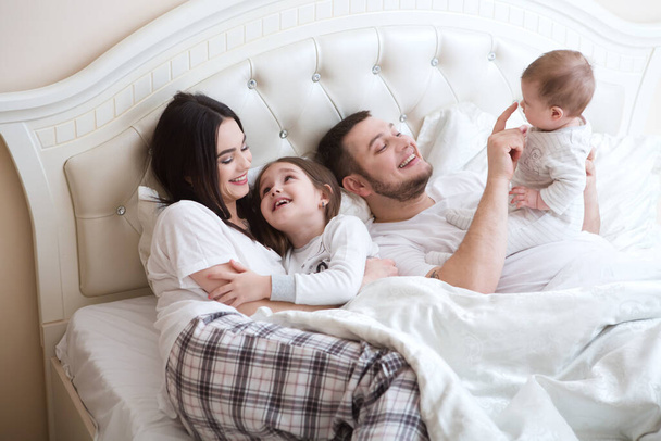 mladí šťastní rodiče s dcerami, kteří si spolu hrají v posteli, rodinný koncept  - Fotografie, Obrázek