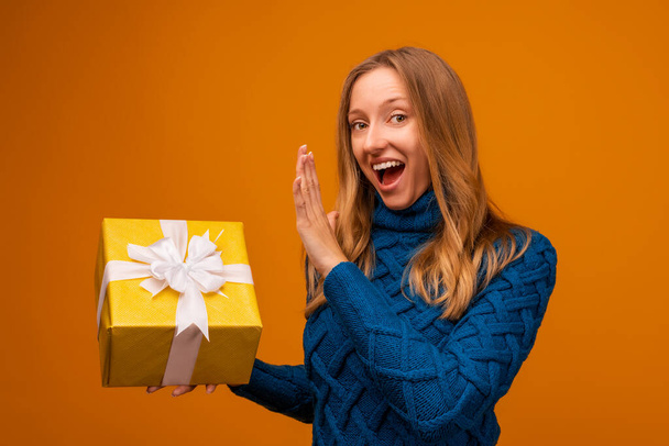 Portrait of a happy young woman in knitted blue sweater holding gift decorated with ribbon. Studio shot, yellow background. New Year, Women's Day, Birthday, Holiday concept - Φωτογραφία, εικόνα