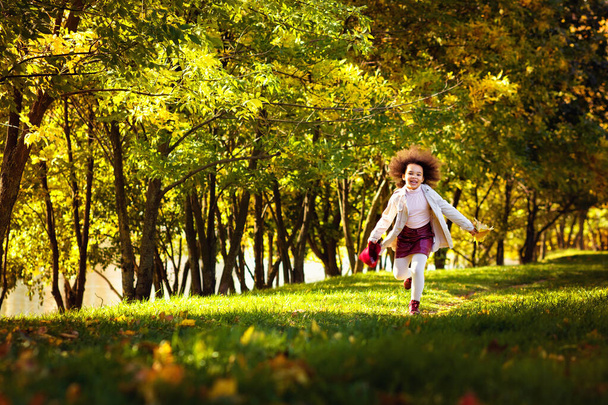 Dziewczyna bawiąca się opadłymi liśćmi. Urocza uczennica spaceruje po szkole po jesiennym parku. Ciemny - oskórowany dziecko rzucam jesienny liść. Dziecięca moda. Jesienne wakacje, styl życia. Szczęśliwego dzieciństwa - Zdjęcie, obraz
