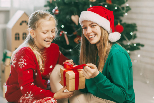 Δύο χαρούμενα και όμορφα έφηβα κορίτσια με ρούχα Πρωτοχρονιάς ανοίγουν δώρα στο σπίτι κάτω από το χριστουγεννιάτικο δέντρο. Χριστουγεννιάτικη διάθεση. Έννοια για τις διακοπές της Πρωτοχρονιάς στο σπίτι - Φωτογραφία, εικόνα