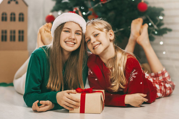 Dos hermanas alegres y alegres en los trajes a la moda de Año Nuevo abren los regalos en casa bajo el árbol de Navidad. Humor de Navidad. Concepto para vacaciones de Año Nuevo en casa - Foto, Imagen