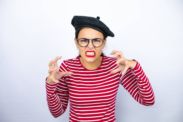 Young beautiful brunette woman wearing french beret and glasses over white background crazy and mad shouting and yelling with aggressive expression and arms raised. Frustration concept. - Photo, Image