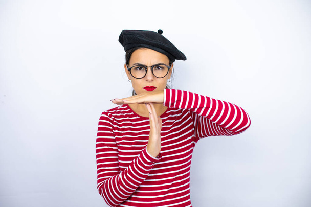 Giovane bella donna bruna con berretto francese e occhiali su sfondo bianco Fare il gesto del tempo con le mani, viso frustrato e serio - Foto, immagini