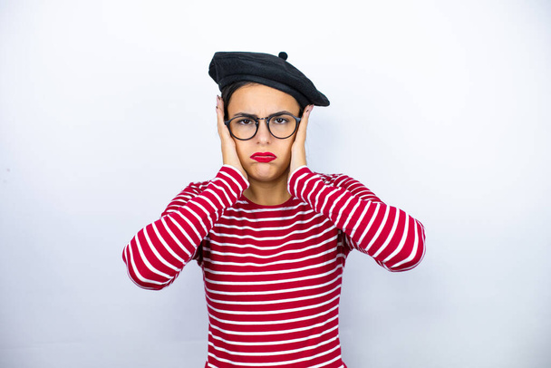 Junge schöne brünette Frau mit französischer Baskenmütze und Brille vor weißem Hintergrund denkt müde und gelangweilt mit den Händen auf dem Kopf - Foto, Bild