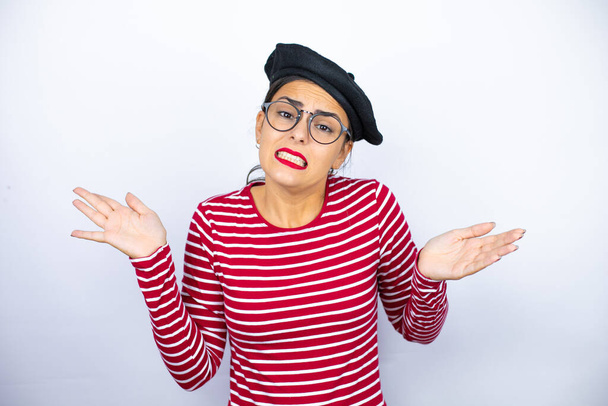 Young beautiful brunette woman wearing french beret and glasses over white background clueless and confused expression with arms and hands raised - Photo, Image
