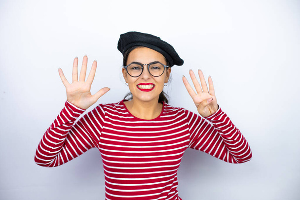 Mladá krásná brunetka žena nosí francouzský baret a brýle nad bílým pozadím ukazuje a ukazuje s prsty číslo devět a zároveň se usmívá sebevědomě a šťastně - Fotografie, Obrázek