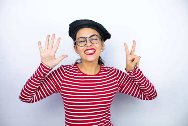 Mladá krásná brunetka žena nosí francouzský baret a brýle nad bílým pozadím ukazuje a ukazuje s prsty číslo sedm a zároveň se usmívá sebevědomě a šťastně - Fotografie, Obrázek