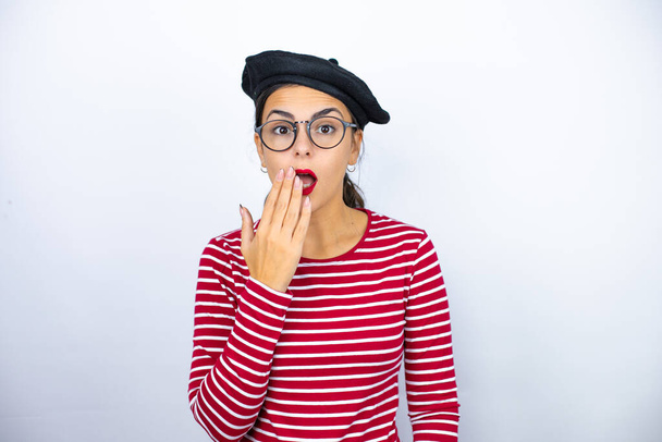 Junge schöne brünette Frau trägt französische Baskenmütze und Brille über weißem Hintergrund überrascht bedeckt ihren Mund - Foto, Bild
