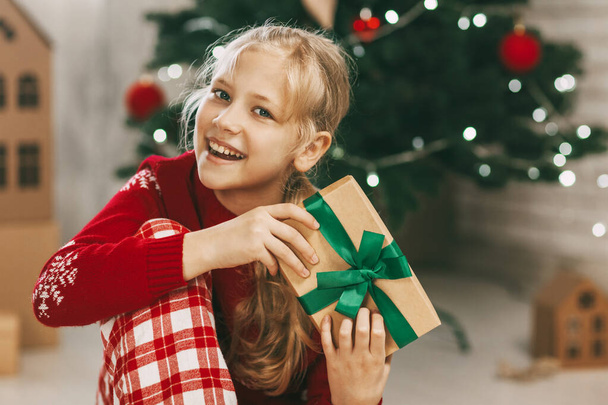 Kaunis pikkutyttö katsoo kameraan ja hymyilee pitäen kädessään vihreällä nauhalla sidottua voimapaperilahjaa. Joulutunnelmaa. Konsepti uudenvuoden loma kotona. - Valokuva, kuva