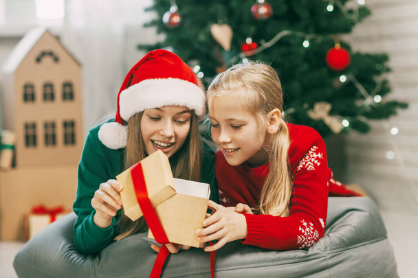 新年の服を着た2人の幸せで美しい10代の女の子がクリスマスツリーの下で家で贈り物を開きます。クリスマス気分。家での新年の休日の概念. - 写真・画像