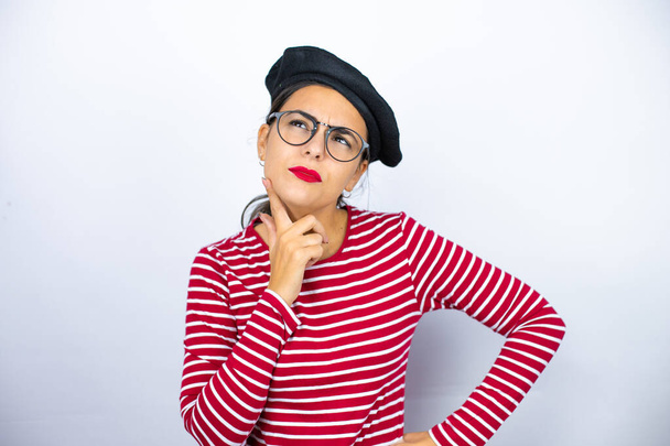Junge schöne brünette Frau mit französischer Baskenmütze und Brille über weißem Hintergrund denken und zur Seite schauen - Foto, Bild