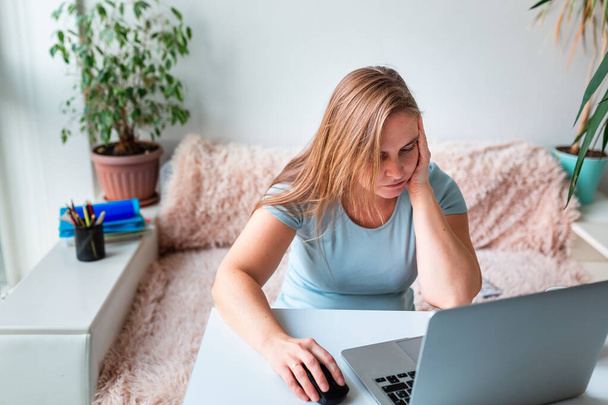 Женщина средних лет устала и ей скучно сидеть за столом дома, работая на ноутбуке. Работа из дома и пребывание дома во время пандемии короновируса - Фото, изображение