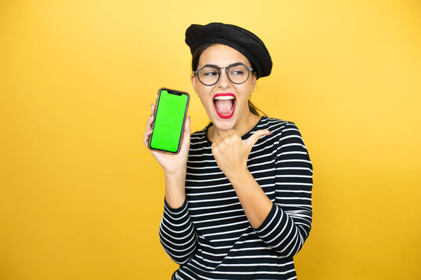 Young beautiful brunette woman wearing french beret and glasses over yellow background smiling, showing the phone and pointing side - Fotografie, Obrázek