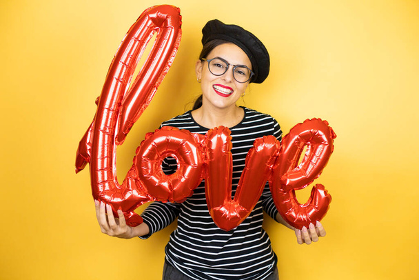 Young beautiful brunette woman wearing french beret and glasses over yellow background holding the word love balloon - Photo, Image