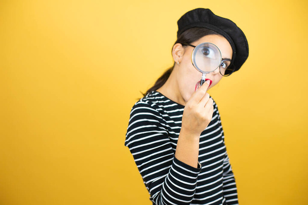 Молодая красивая брюнетка в французском берете и очках на желтом фоне удивлена, глядя через лупу - Фото, изображение