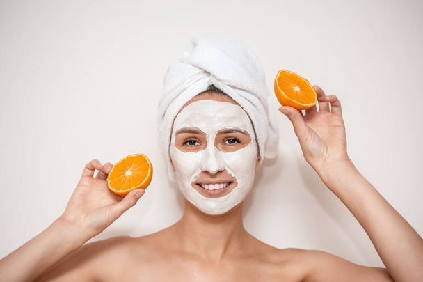 Zbliżenie na piękną uśmiechniętą kobietę z białą maską i pomarańczowym kawałkiem. piękna dziewczyna z ręcznikiem na głowie stosowania maski twarzy. - Zdjęcie, obraz