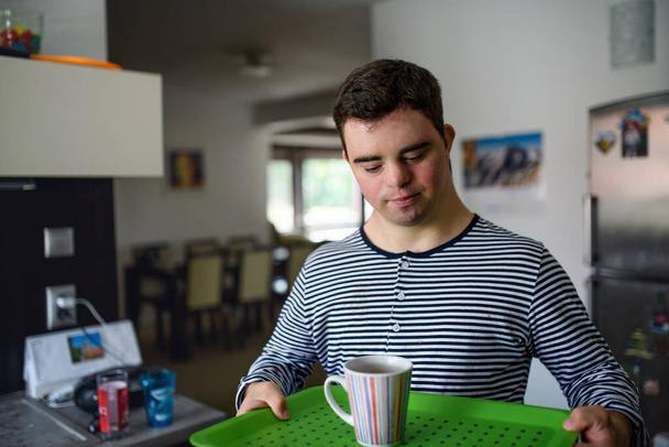 Down syndrome adult man standing indoors in kitchen at home, carrying cup on tray. - Foto, Imagem