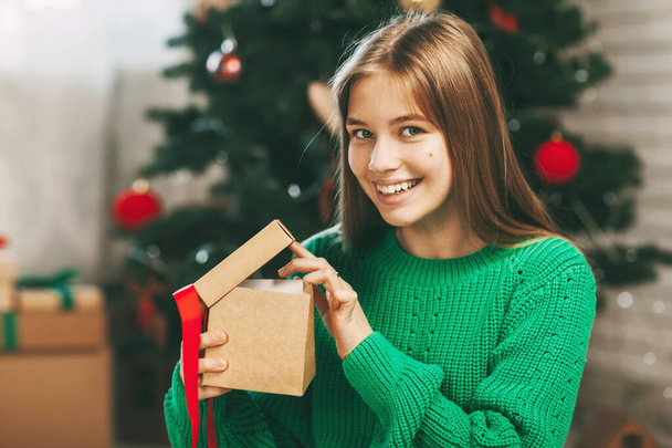 Kaunis teini-ikäinen tyttö avaa lahjan, joka on valmistettu kraft-paperista, joka on sidottu punaisella nauhalla, uudenvuoden tunnelmalla. Joulun ja uudenvuoden käsite kotona - Valokuva, kuva