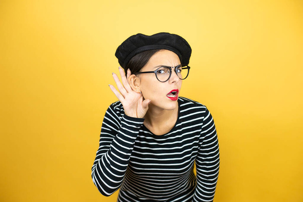 Giovane bella donna bruna con berretto francese e occhiali su sfondo giallo sorpreso con mano sull'orecchio ascoltando un'udienza a pettegolezzi o pettegolezzi - Foto, immagini