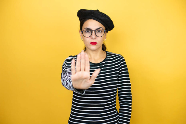 Young beautiful brunette woman wearing french beret and glasses over yellow background serious and doing stop sing with palm of the hand. - Photo, Image