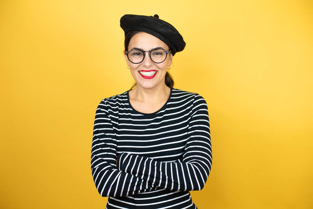 Young beautiful brunette woman wearing french beret and glasses over yellow background with angry face, negative sign showing dislike with thumbs down - Photo, image