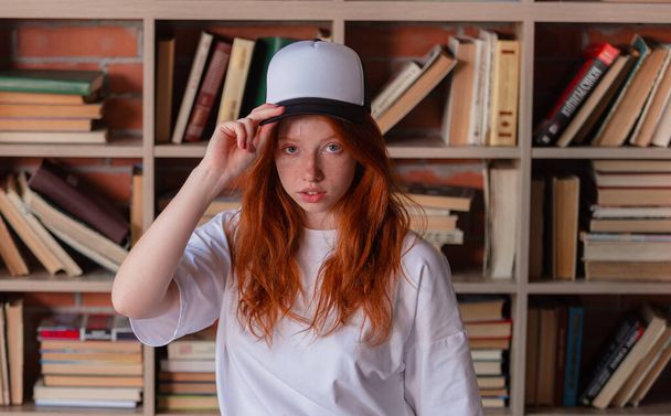 Portrait of a redhead woman in template blank t-shirt and cap with healthy freckled skin. Young caucasian model posing on bookshelf background. Copy space and mock up. Place for adverising - Photo, Image