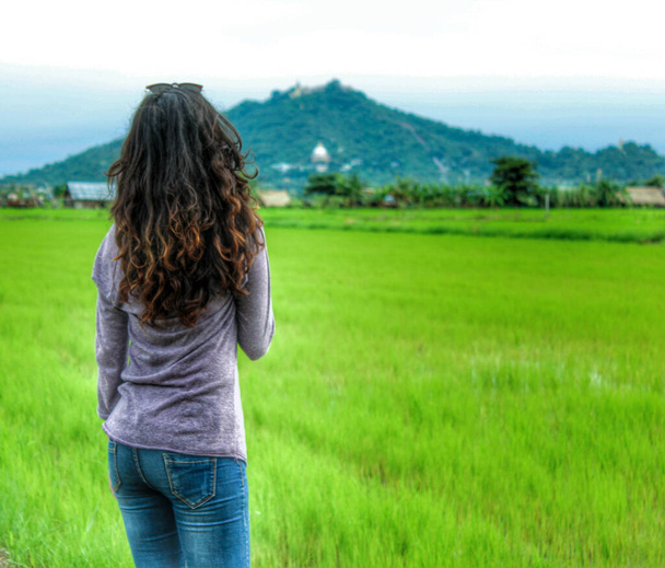 Όμορφο κορίτσι με σγουρά μαλλιά βλέποντας πράσινο πεδίο και βουνό από πίσω - Φωτογραφία, εικόνα