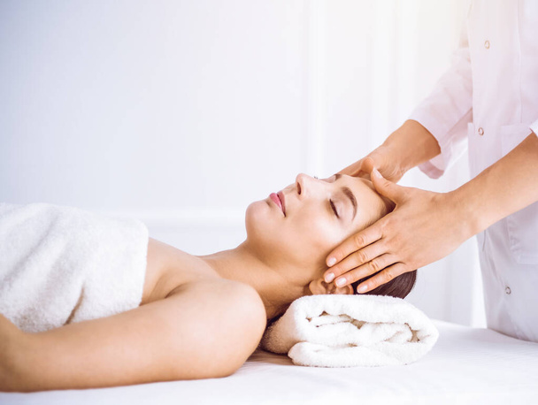 Beautiful brunette woman enjoying facial massage with closed eyes in sunny spa center . Relaxing treatment and cosmetic medicine concepts - Fotoğraf, Görsel