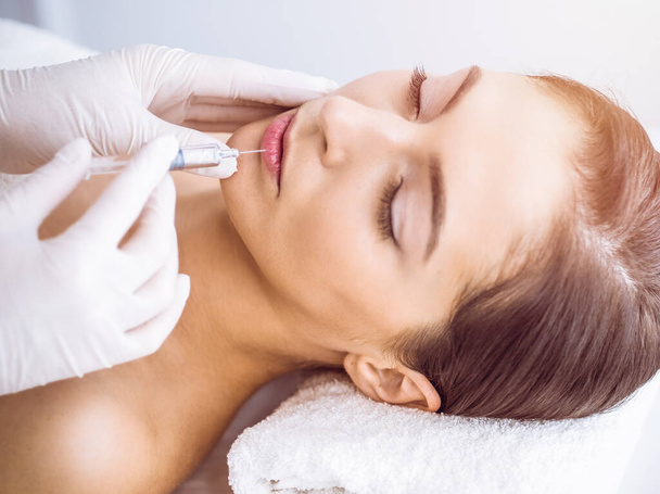 Beautician doing beauty procedure with syringe to face of young brunette woman in sunny spa center. Cosmetic medicine and surgery, beauty injections - Fotografie, Obrázek