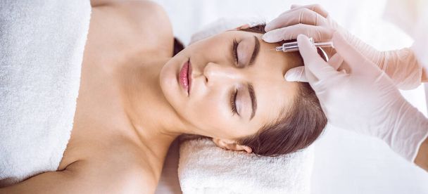 Beautician doing beauty procedure with syringe to face of young brunette woman in sunny spa center. Cosmetic medicine and surgery, beauty injections - Fotó, kép