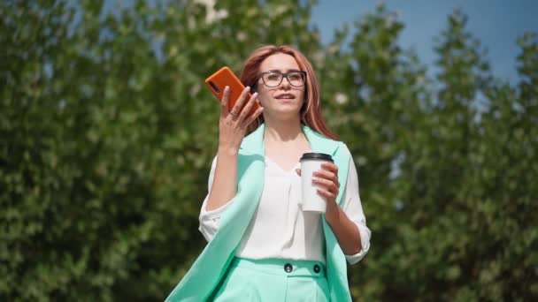 Beautiful business woman in glasses and a trendy turquoise suit with paper cup coffee in her hands walk and records voice message on mobile phone in downtown. Pretty lady uses smartphone. Slow motion. - Footage, Video