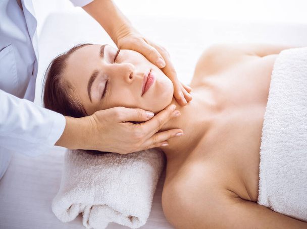 Beautiful brunette woman enjoying facial massage with closed eyes in sunny spa center . Relaxing treatment and cosmetic medicine concepts - Foto, Imagem