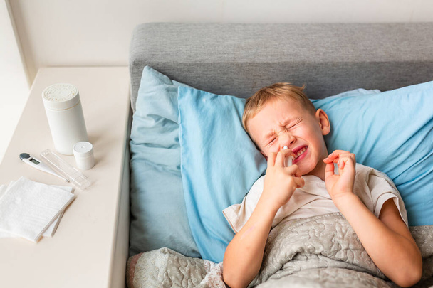 Sick little boy with high fever and headache laying in bed spraying medicine into his nose. Stay at home during corona virus epidemic if you feel sick - Foto, Imagem
