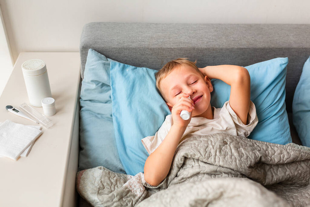 Sick little boy with high fever and headache laying in bed spraying medicine into his nose. Stay at home during corona virus epidemic if you feel sick - Foto, imagen