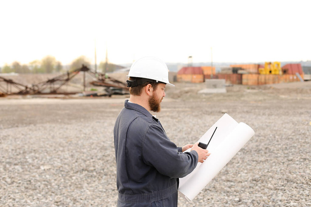 Обратный вид инженера-строителя, стоящего на строительной площадке, разговаривающего по рации УКВ и держащего чертежи. - Фото, изображение