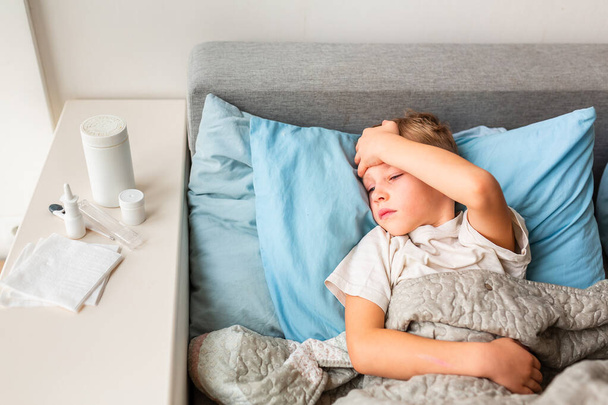 Sick little boy with high fever and headache laying in bed checking his forehead. Stay at home during corona virus epidemic if you feel sick - Valokuva, kuva