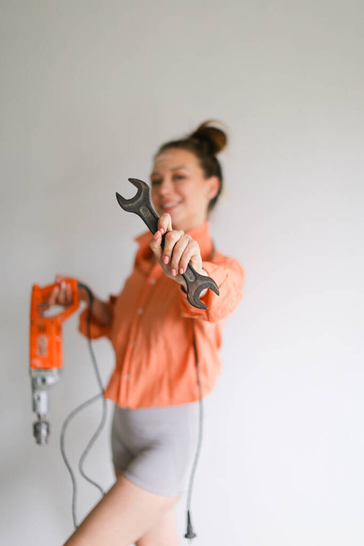 Focus on wrench, young girl holding drill and wearing orange shirt, isolated background. - Foto, Imagen