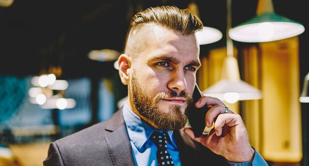 Bearded entrepreneur dressed in formal wear calling via smartphone device for talking about business plan, successful male proud ceo communicating via digital cellular during worktime in office - Fotoğraf, Görsel