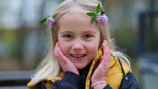 Egy bájos kislány portréja, lóherevirággal a hajában. aranyos lány 5 éves ravasz szem néz félre egy mosollyal nézz a kamerába. Lezárjuk a videót. Lassított felvétel - Felvétel, videó