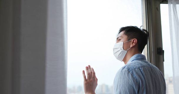 asian young man wearing face mask stay isolation at home for self quarantine due to epidemic of COVID19 - he looks out the window - Foto, Imagen