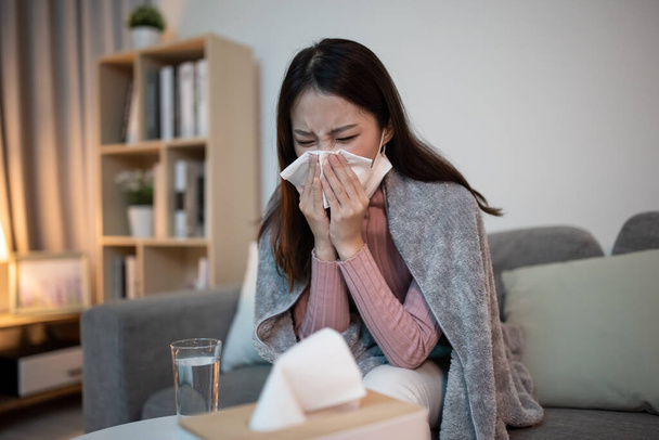 Sick asian woman is blowing running nose and sneezing in tissue at home - Valokuva, kuva