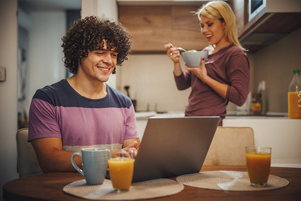 Shot of a happy couple spending their morning time together and talking in the kitchen. They are using laptop and eating breakfast before work. - Photo, Image