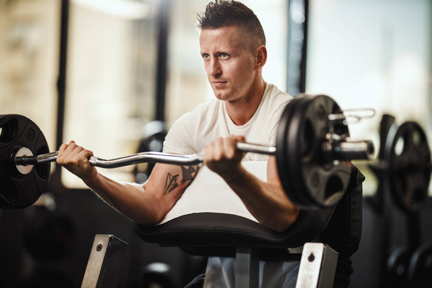 Una foto de un tipo musculoso en ropa deportiva haciendo ejercicio en el duro entrenamiento del gimnasio. Él está bombeando músculo bíceps con peso pesado. - Foto, imagen