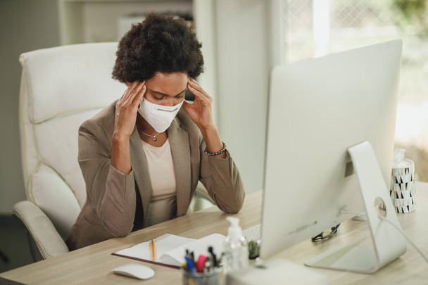 Fotografía de una mujer de negocios africana cansada con máscara protectora N95 sentada sola en su oficina y trabajando en la computadora durante la pandemia de COVID-19. - Foto, imagen