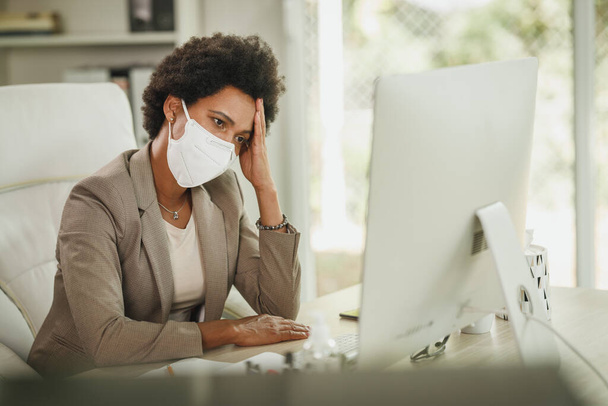 Shot of a worried African businesswoman with protective N95 mask sitting alone in her home office and working on computer during COVID-19 pandemic. - Fotografie, Obrázek